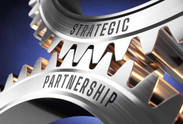 Close-up view of two polished stainless steel gears meshing together, with one gear labeled "STRATEGIC" and the other labeled "PARTNERSHIP," symbolizing business collaboration and operational synergy.