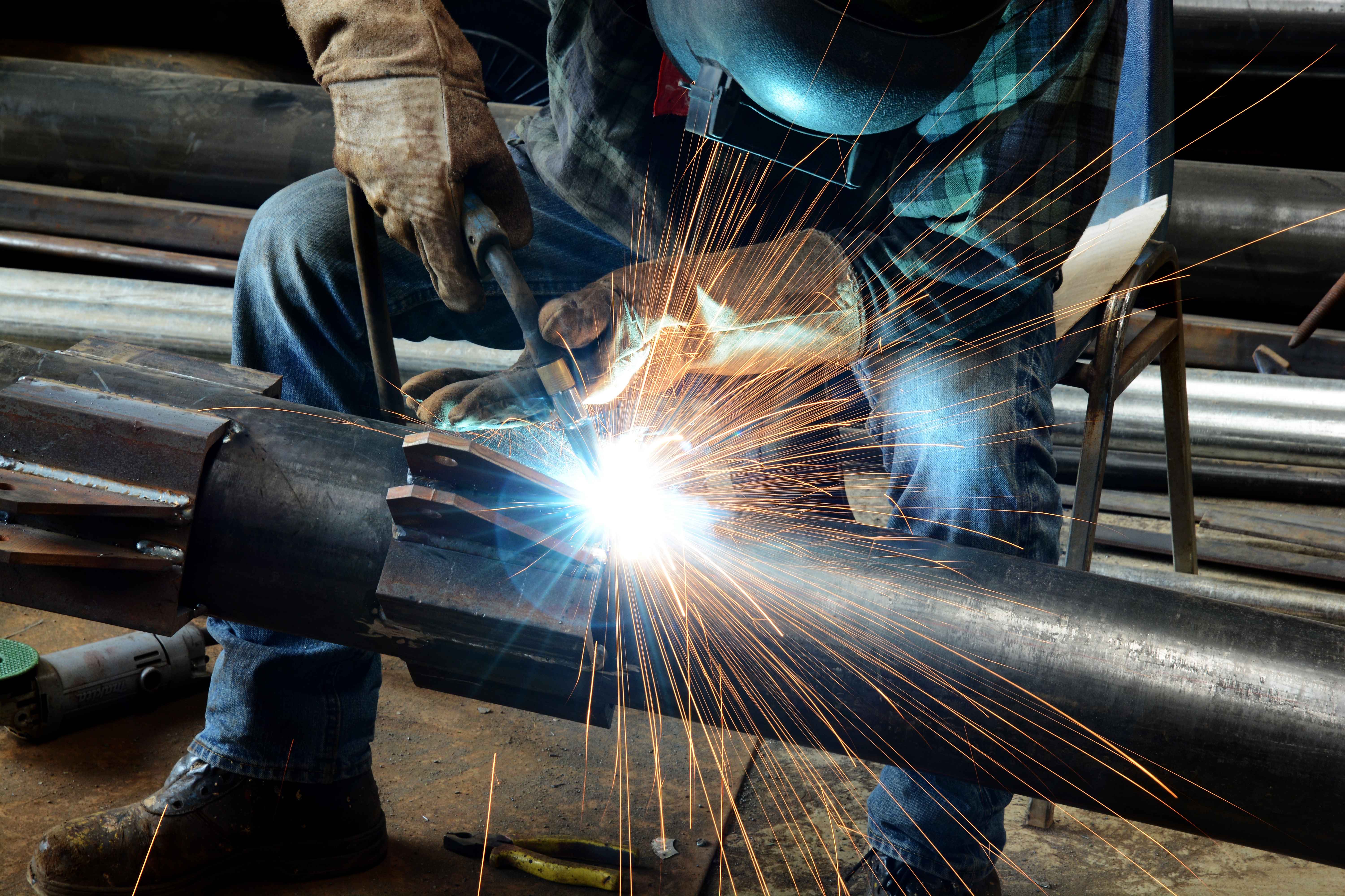 Close-up of a welder joining steel pipe sections in a fabrication workshop, with bright welding sparks radiating from the weld point and metal stock visible in the background.