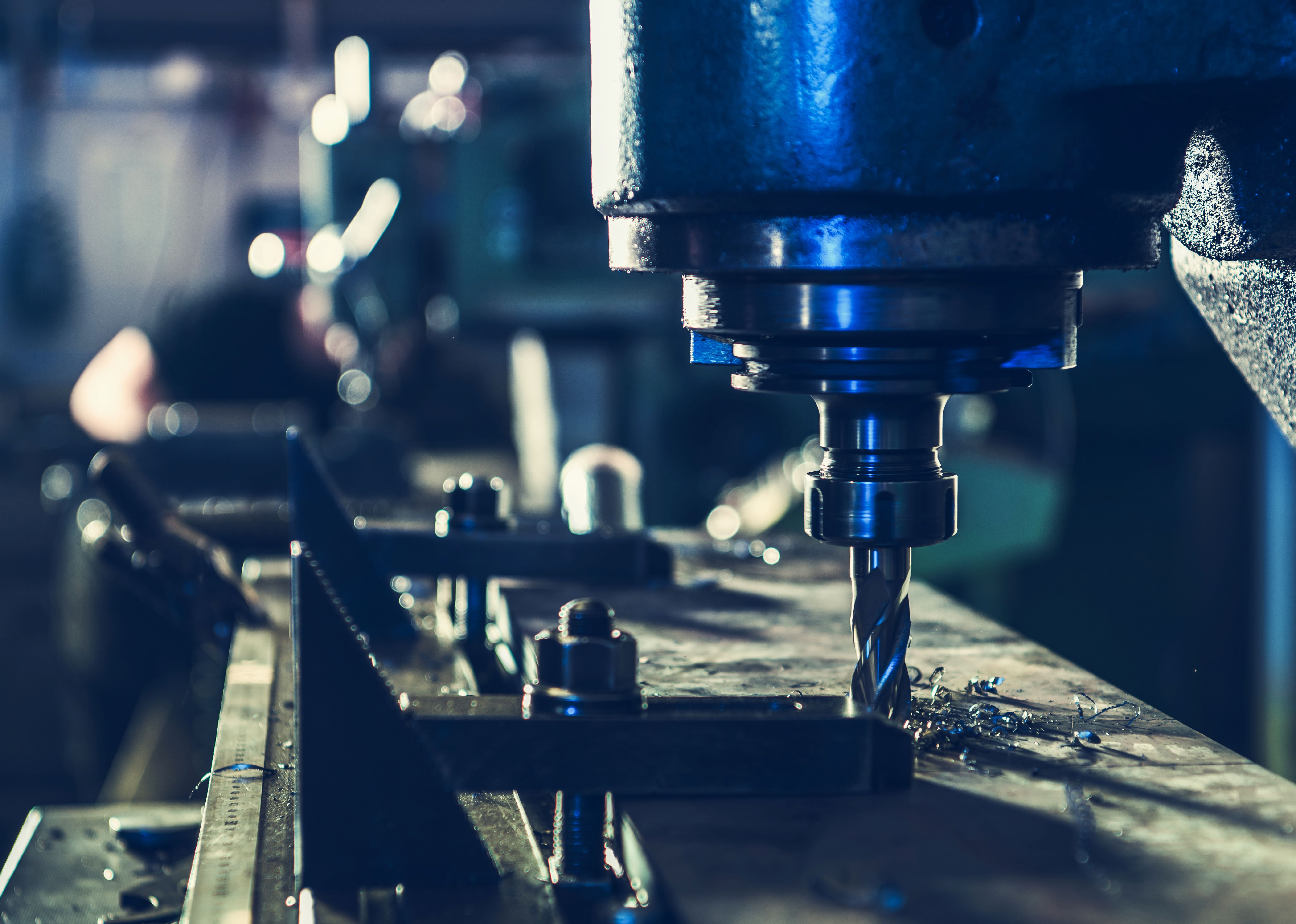 Industrial CNC milling machine cutting a metal workpiece, with a rotating tool and metal shavings visible on the machining surface under blue-toned factory lighting.