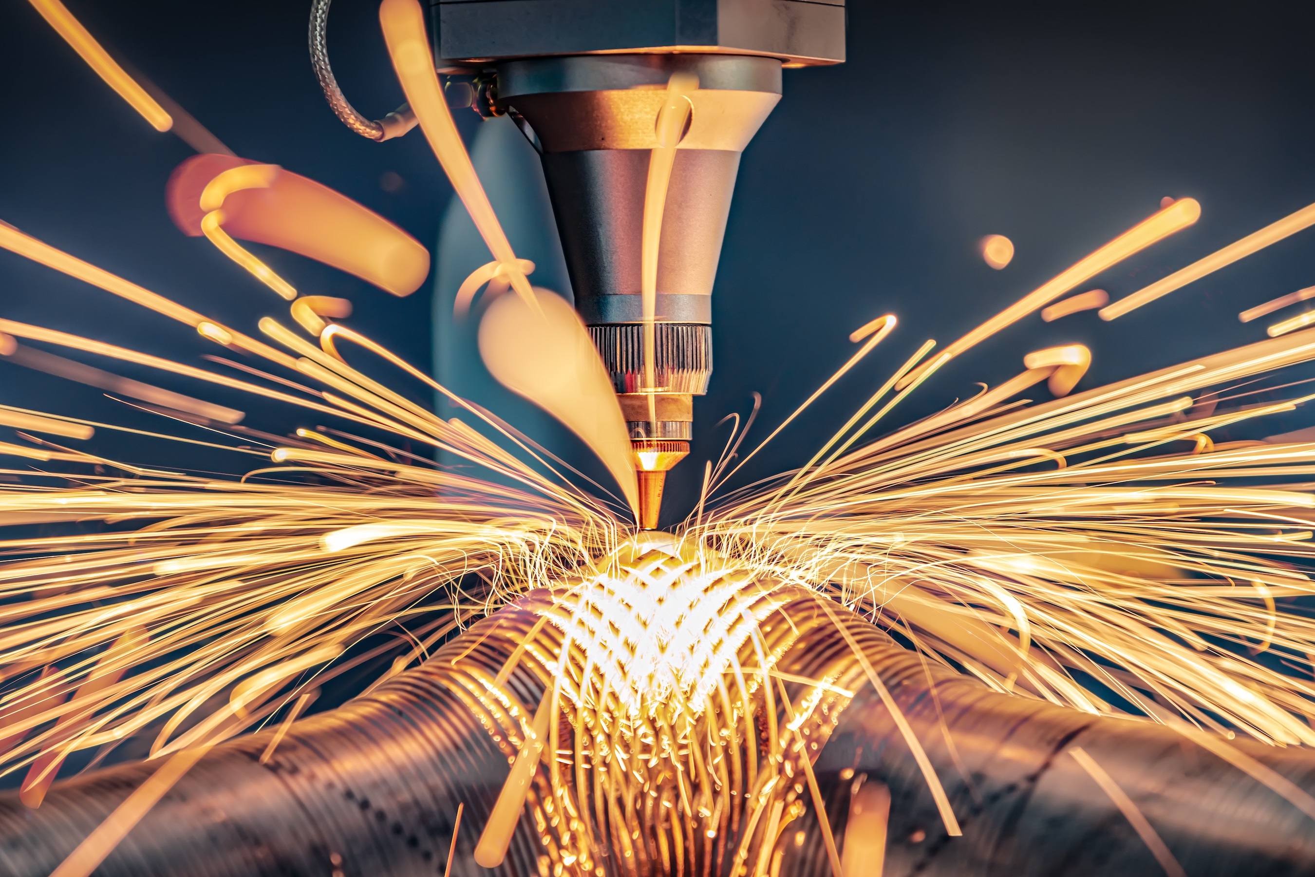 High-precision laser cutting operation on a metal workpiece, with intense orange sparks radiating outward from the cutting point beneath the laser nozzle in a modern manufacturing environment.